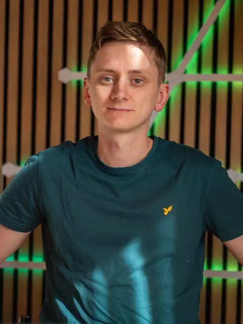 Nils Lorenzen im petrolfarbenen T-Shirt vor einer Holzlamellenwand mit grünem LED-Licht